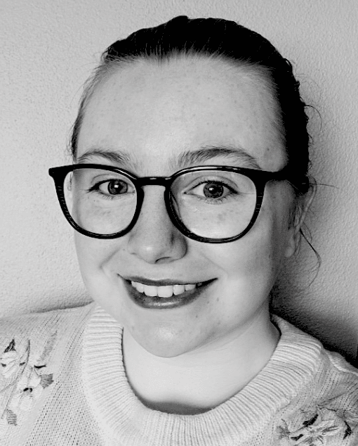 A white woman with glasses and a thoughtful expression leans on a doorframe.