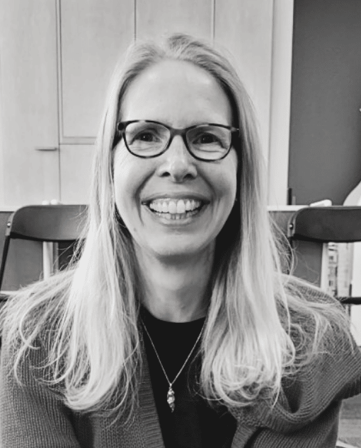 A white woman with glasses and a thoughtful expression leans on a doorframe.